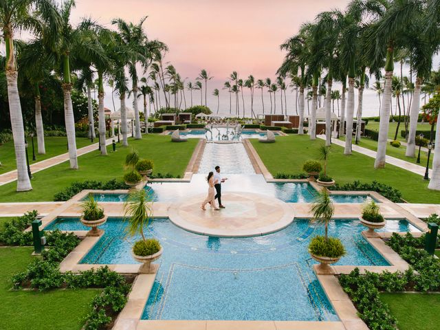 A couple walks on a path across pools and fountains with manicured hotel grounds next to the ocean.
