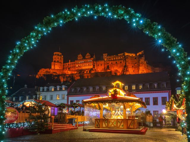A view of the Christmas Market in Heidelberg through a green archway, looking out onto the market square, with the castle in the distance above, at night.