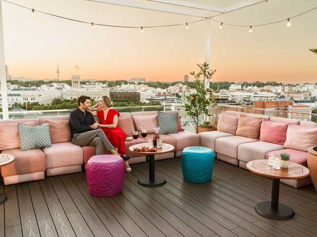 A couple sits on an outdoor sofa drinking sangria atop the Atico rooftop bar with a view of the Madrid skyline at dusk in the background.