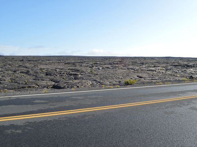 A road with a wide stretch of lava field with cracked black earth and small shrubs.