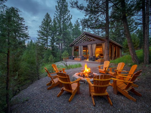 A cabin on a hill in the woods with a fire pit and Adirondack chairs in front of it.