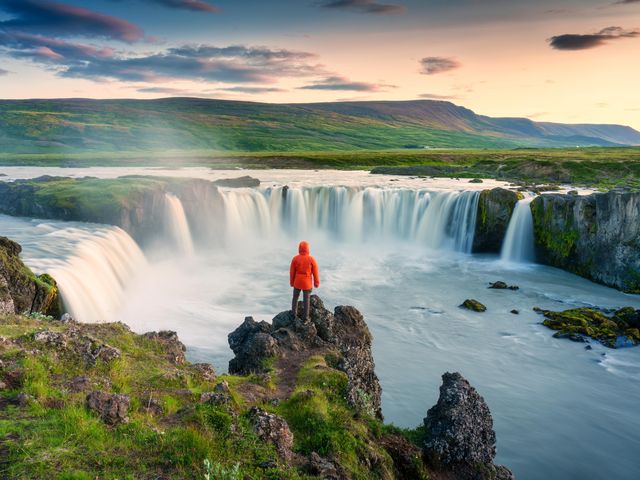A person wearing a red-hooded coat stands on a cliff overlooking a waterfall in Southern Iceland.