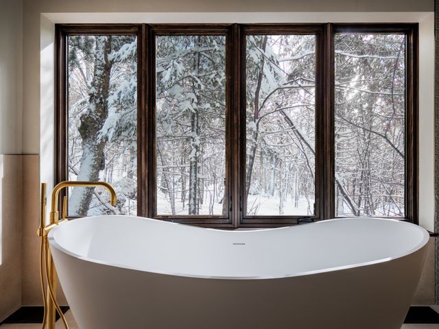 A white soaking bathtub facing large floor to ceiling windows with views of snowy trees. 