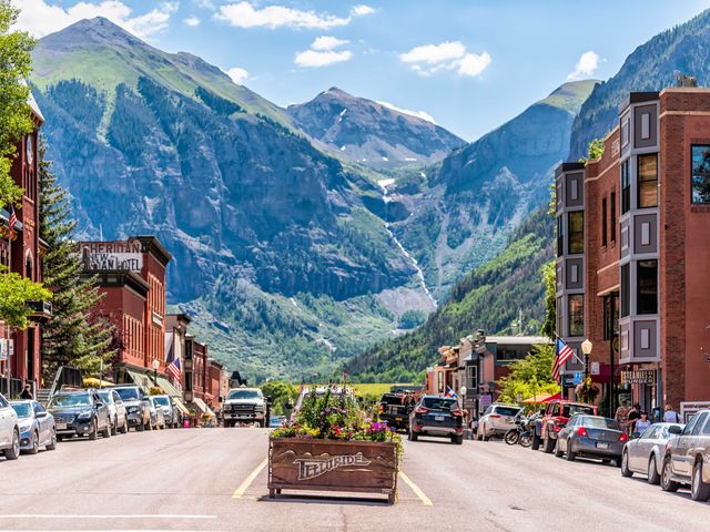 A quaint downtown area with brick buildings on either side surrounded by green mountain views and a Telluride sign in the middle.