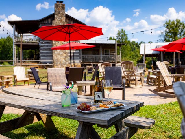 A picnic table in a winery with drinks and food on a patio with Adirondack chairs and red umbrellas. 