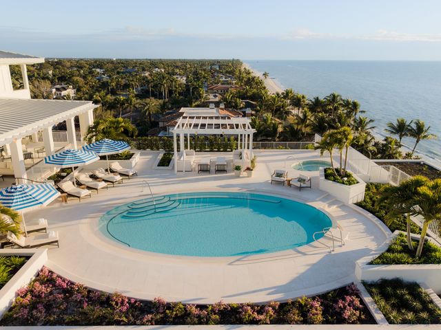 An oval-shaped pool on a terrace facing the ocean with blue-and-white umbrellas and white pool chairs. 