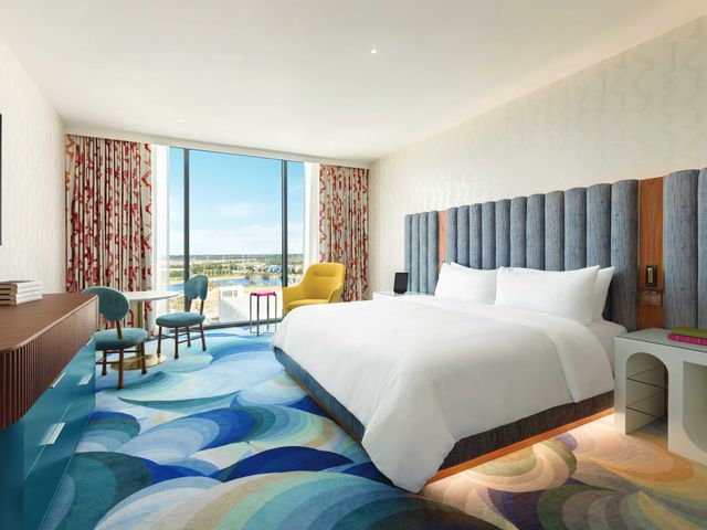 A hotel guest room with a large bed, modern carpeting, large windows, and a plush headboard.