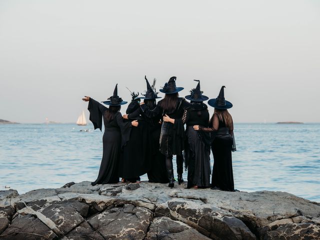 A group of women dressed as witches pose at the beach with their arms around each other, as seen from behind.