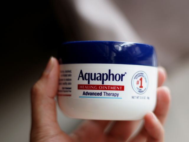 A hand holding a small tub of Aquaphor healing ointment.
