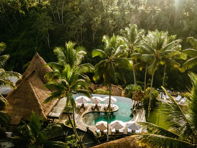 A turquoise infinity pool surrounded by a thatched roof building and lush green trees at Viceroy Bali.
