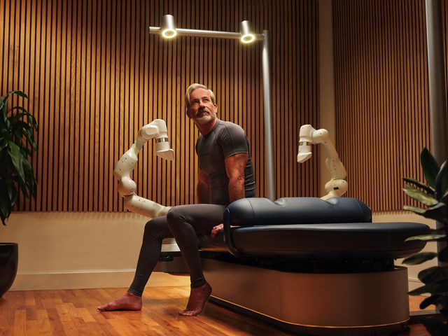 A man sits on an Aescape massage table with its white robot arms visible in a wood paneled room.