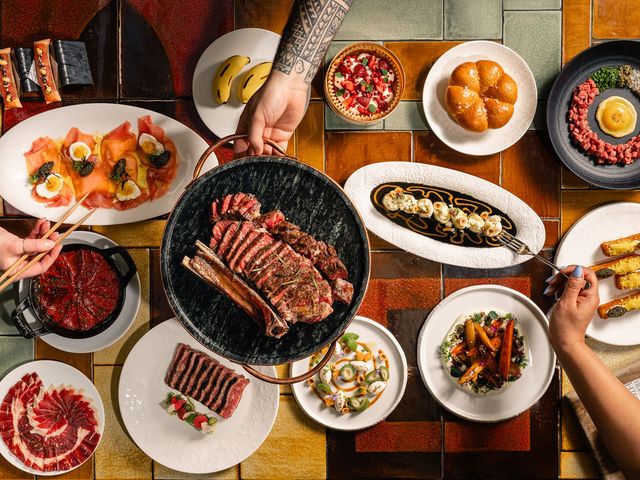 An overhead shot of a table packed with dishes of food and one hand golding chopsticks, another a fork, and one holding a pan full of meats.