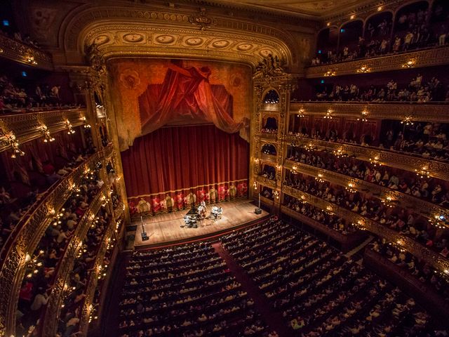 A full theatre packed with balconies on the side and red velvet curtains on the stage.