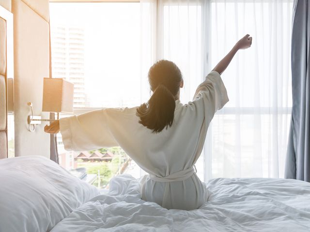 A woman as seen from the back in a hotel robe on a bed stretching while looking out the window.