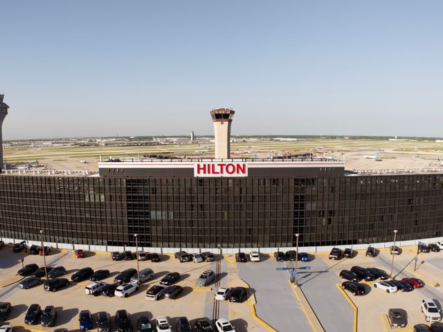 A large airport building that says Hilton surrounded by parked cars and air traffic control towers.