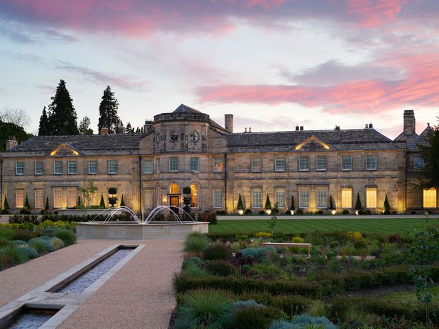 The exterior stone estate of Grantley Hall is lit up at dusk with a fountain in front of it and landscaped areas.