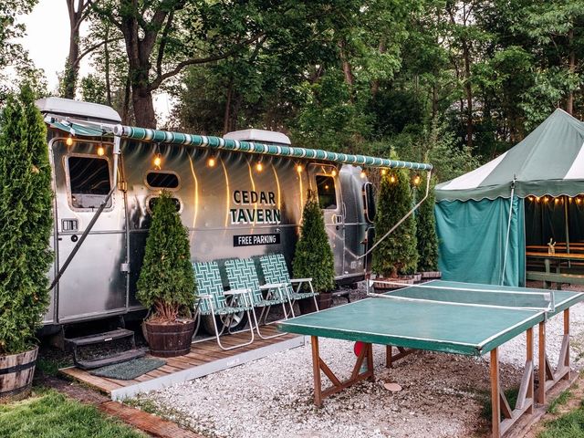 A parked Airstream trailer with a tavern sign on it, with a ping pong table and nearby picnic tables in a tent.