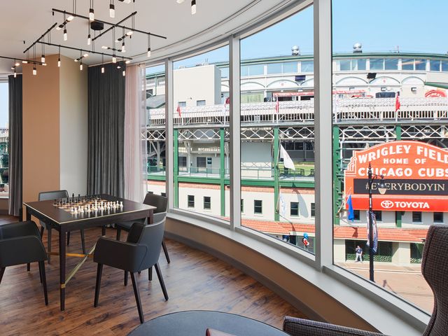 A hotel room with a chess table next to floor-to-ceiling windows with views of the entrance to Wrigley Field.