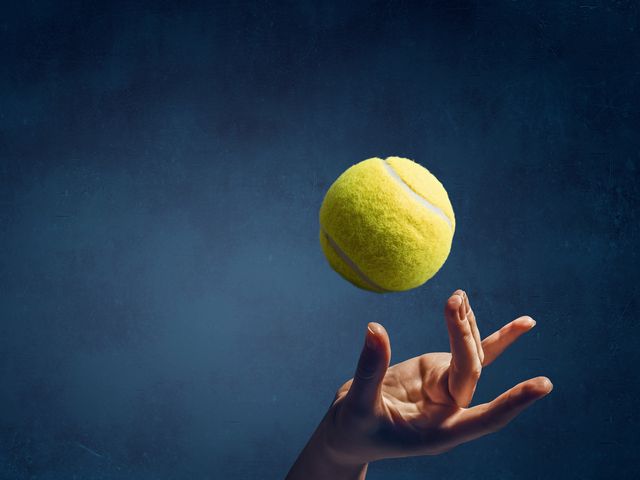 A hand tossing a tennis ball in the air against a dark background.