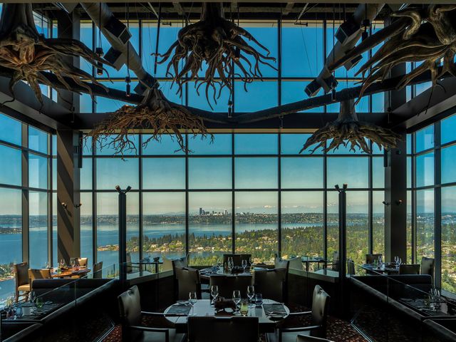 An empty dining room with rustic twisted wood chandeliers and large windows overlooking a city on the water.
