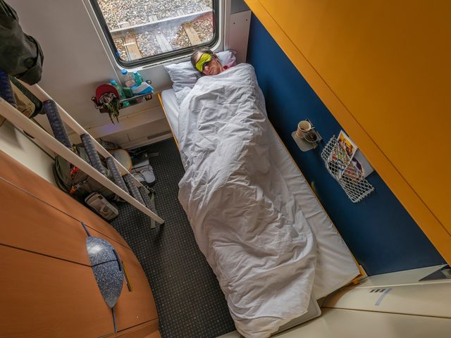 A person is asleep on a sleeper train car wrapped up in white blankets in a small room.