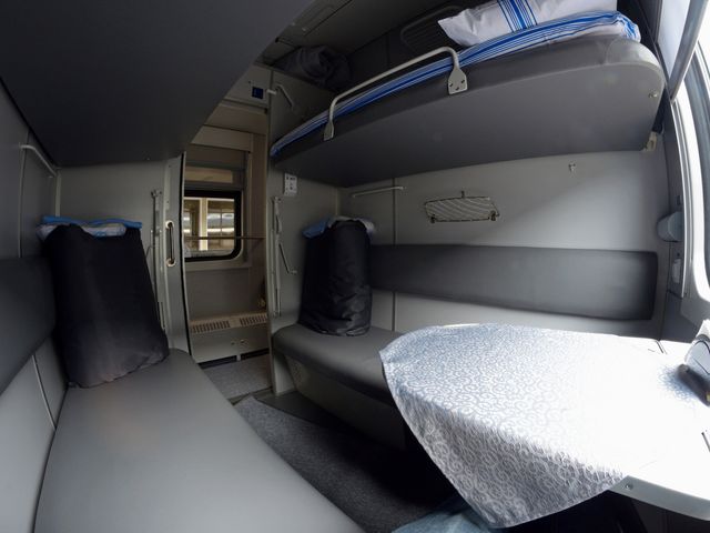 The interior of a sleeper train, with a simple twin bunk bed, a couch, and gray decor.