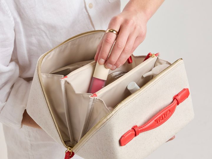 A hand reaching into the Calpak lay flat toiletry bag while open to put in lipstick.