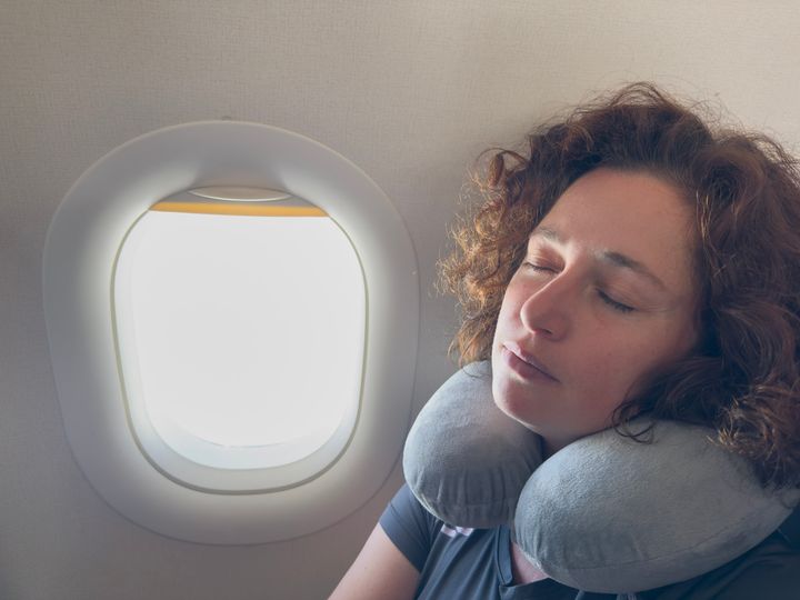 A woman with curly hair sleeping next to a plane window wearing a neck pillow.