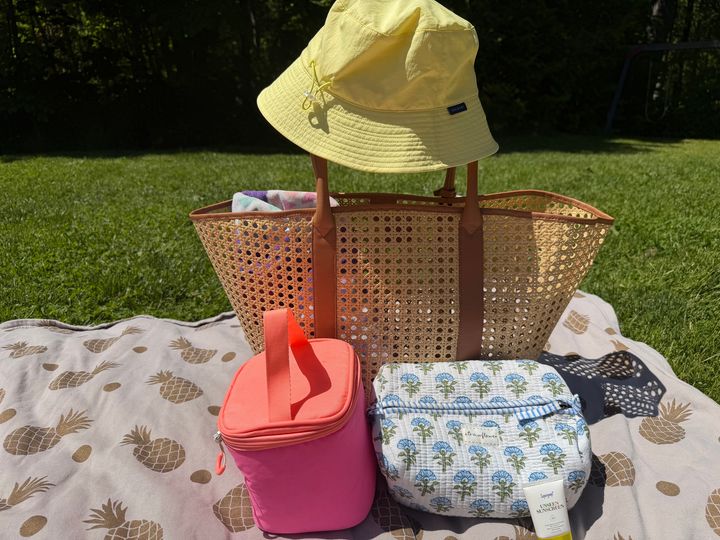 A beach bag on a towel with a hat on the handle, cooler bag, and a toiletry bag.