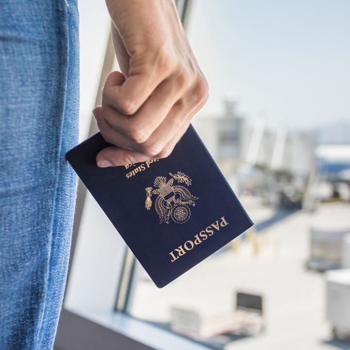 A hand is holding a USA passport at the airport next to a window with a plane visible.