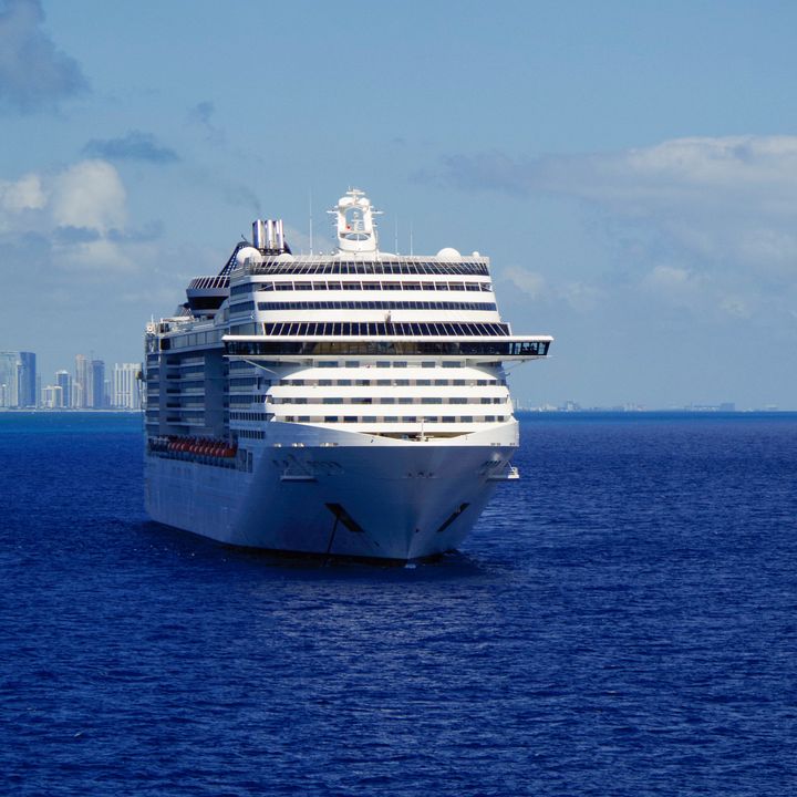 Two cruise ships sit at sea with skyscrapers far in the distance at a port.