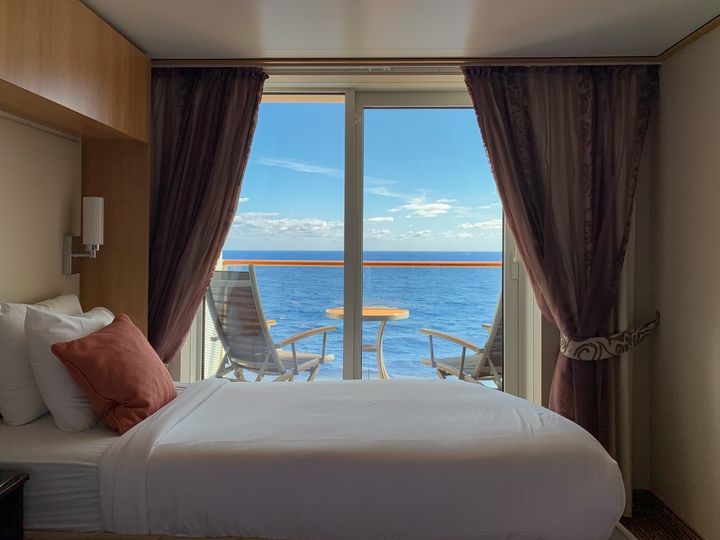A cruise ship stateroom with a bed next to a balcony with two chairs overlooking the blue ocean.