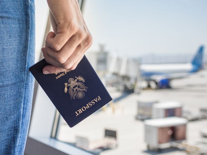 A hand is holding a USA passport at the airport next to a window with a plane visible.  