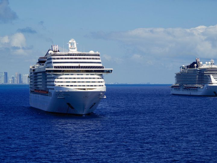 Two cruise ships sit at sea with skyscrapers far in the distance at a port.