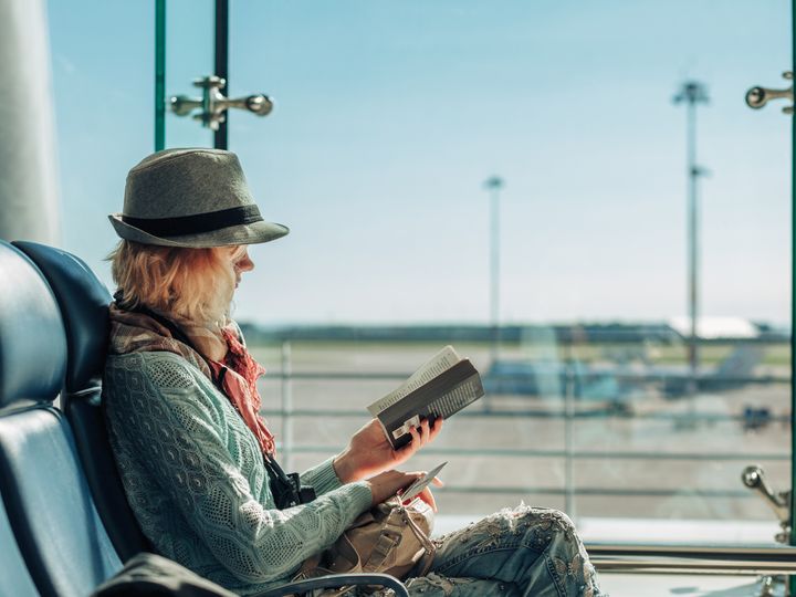 Female traveler wearing hat, scarf, sweater, and jeans reads a paperback at an airport gate.