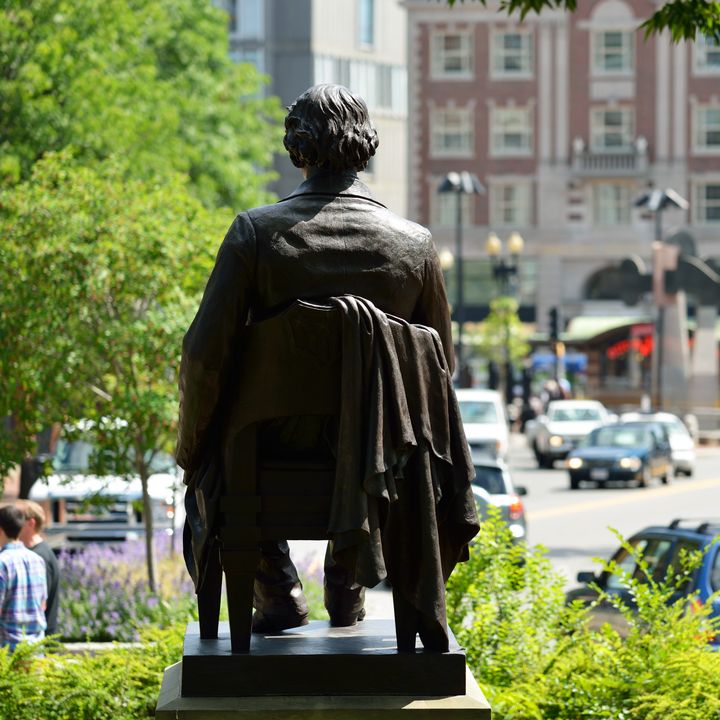 John Harvard Statue in Harvard Square as seen overlooking traffic and buildings.