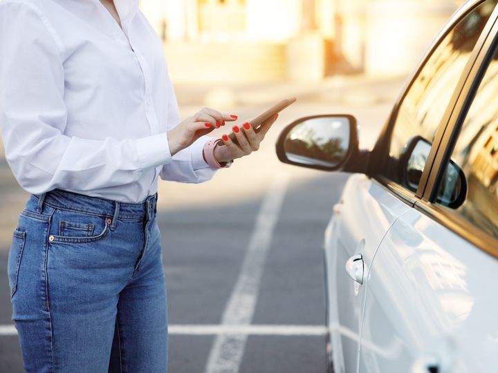 A woman from the neck down is seen on her phone next to a car.