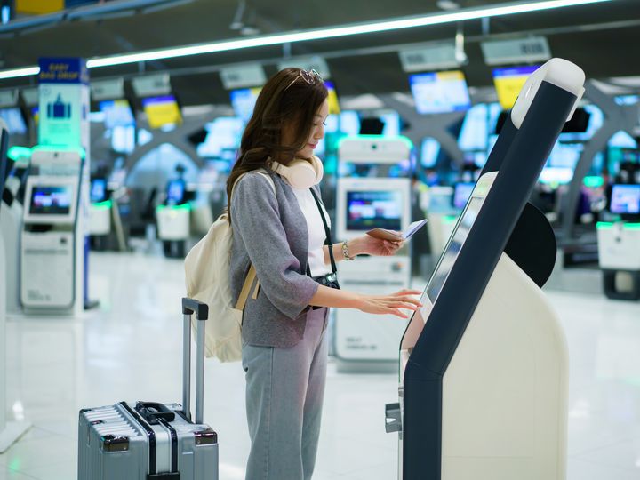 A woman with headphones around her neck and a suitcase at her side stands and operates an airport check-in kiosk