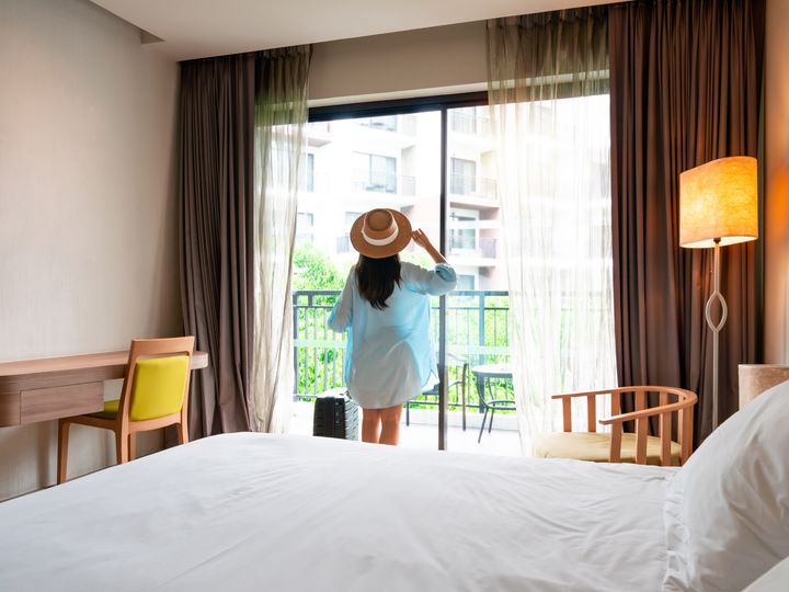 A woman looking out the window of a hotel room wearing a floppy hat and holding her suitcase next to the bed.