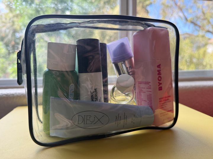 A clear Packism TSA-friendly travel bag filled with products on a table in front of a window.