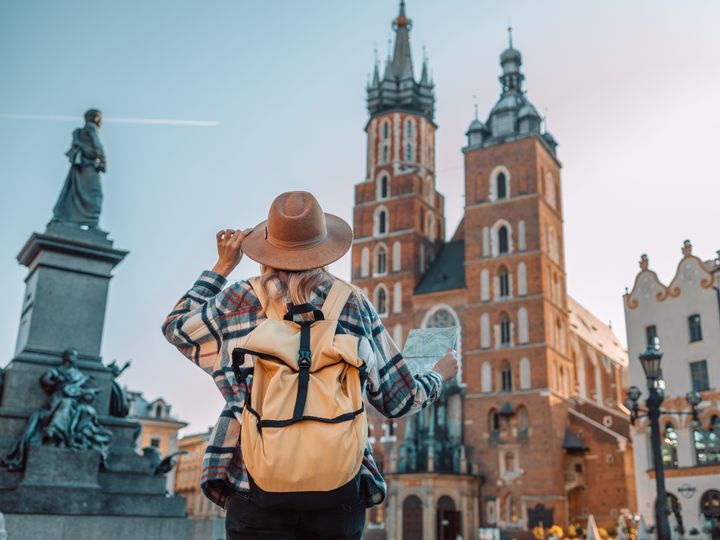 A tourist in Europe wearing a hat and a backpack as seen from behind next to historic buildings.