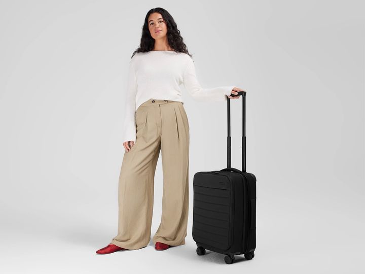 A woman stands against a neutral background holding the handle to an Away wheeled soft-sided carry-on.