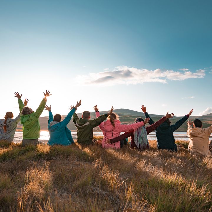 A group of people sits with their backs to the camera and arms in the air in a scenic setting at sunset.
