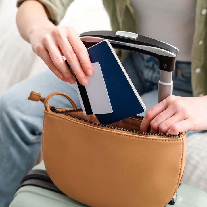 A person is seen without the face visible putting a passport and credit card into a small purse on top of luggage.