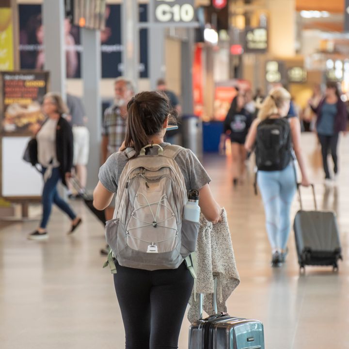 A woman is seen from behind wearing a backpack and pulling her suitcase while walking next to airport shops.