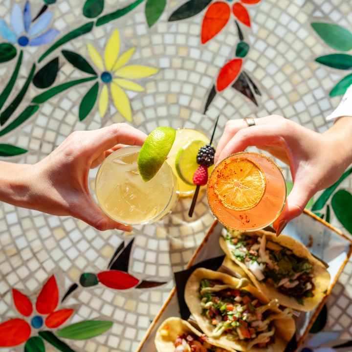 An overhead view of two sets of hands clinking cocktails over a mosaic tiled-table with tacos.