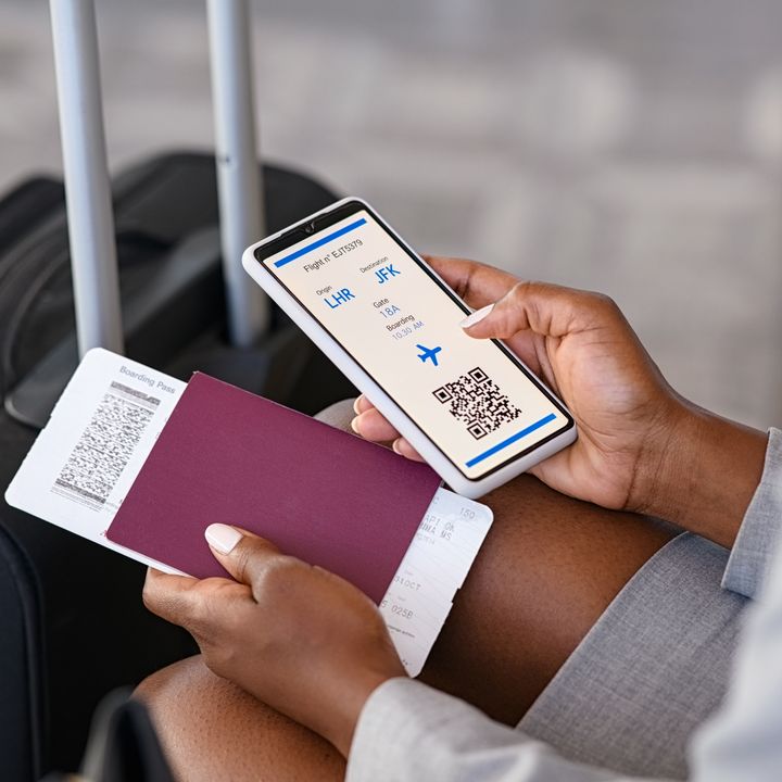 A woman’s hand is seen holding a smart phone with her digital boarding pass visible on her app, next to her passport in her other hand and luggage.