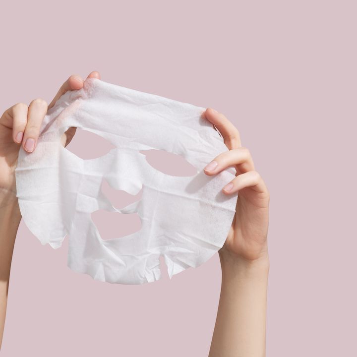 Female hands holding sheet of white mask on pink background.
