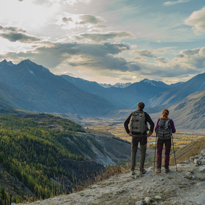 A couple of hikers with backpacks standing on mountain peak looking at vast valley, panoramic view of snow capped mountains and autumn forest in Mongolia
