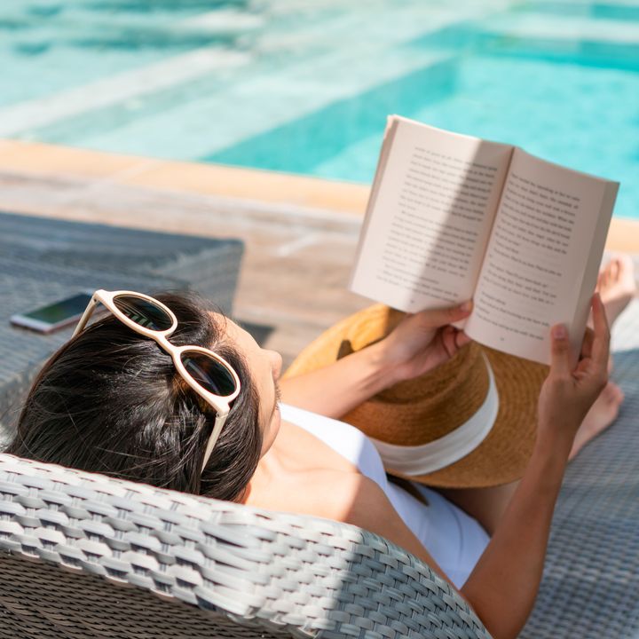 A woman is seen from behind on a pool chair reading a book by the water with her sunglasses on top of her head.
